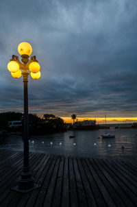 Illuminated street lights by sea against sky during sunset