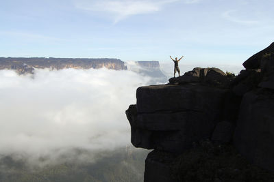 Full length of man with arms raised standing on rocky mountains against sky