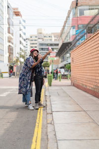 Full body side view of content peruvian female friends taking self portrait on smartphone while standing near road in city