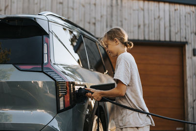 Side view of boy inserting charging plug in electric car on driveway