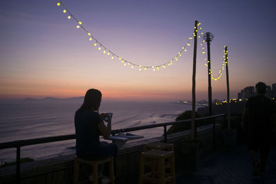 Silhouette woman using mobile while sitting on stool at promenade during sunset