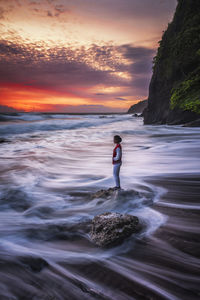 Woman standing on rock at beach against sky during sunset