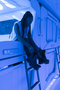 Side view of african american female taking off boots and getting ready for relaxing in room of capsule hotel illuminated by blue light