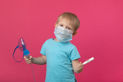 A blond child with an inhaler and a thermometer in his hand on a plain pink background. studio. 