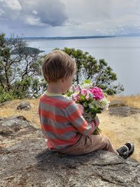 Rear view of boy holding bouquet while sitting on rock against sky