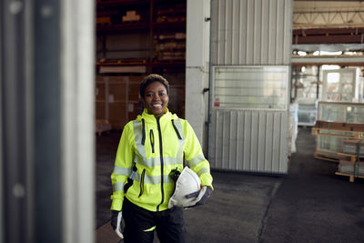 Portrait of smiling blue-collar worker in protective workwear holding hardhat in factory