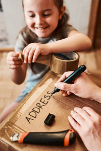 Smiling cute girl holding adhesive tape while cropped hands writing over cardboard paper