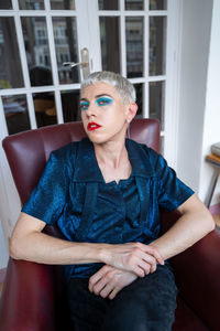 Portrait of confident androgynous man wearing makeup sitting on armchair at home