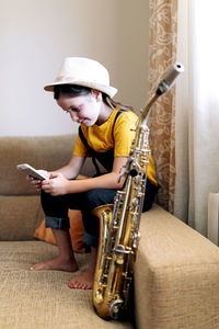 Child in hat text messaging on cellphone while sitting on couch with saxophone in living room