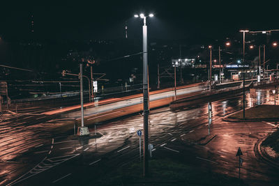 Illuminated railroad tracks by street against sky at night