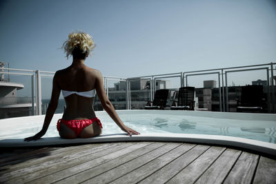 Rear view of beautiful woman sitting at poolside on building terrace against clear sky
