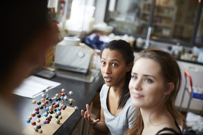 Young female students by molecular structure looking at mature teacher in chemistry classroom