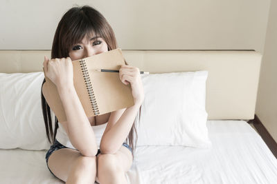 Portrait of young woman holding spiral notebook while sitting on bed