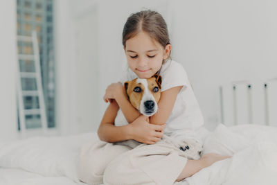 Cute girl embracing dog while sitting on bed at home