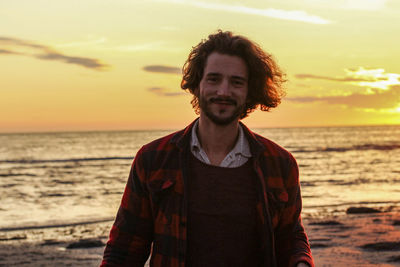 Portrait of smiling young man standing at beach against sky during sunset