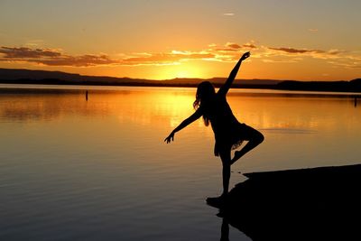 Silhouette woman with arms outstretched by lake against sky during sunset