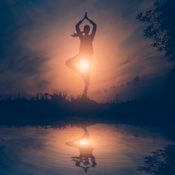 Silhouette woman meditating while standing by lake against sky during sunset