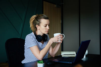 Caucasian woman sitting home office workplace, holds cup of coffee and works with laptop