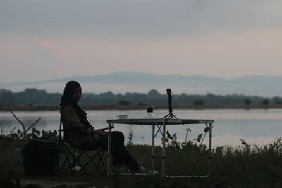 Man sitting on seat by lake against sky during sunset