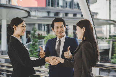 Female colleagues giving handshake while businessman showing thumbs up while standing in office corridor