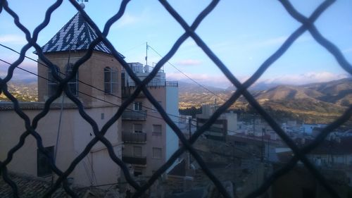 Buildings against sky seen through chainlink fence