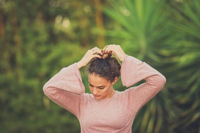 Woman making hair bun while standing against trees