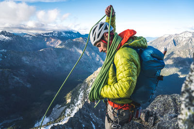 Man wearing jacket and backpack coiling rope on rock climb