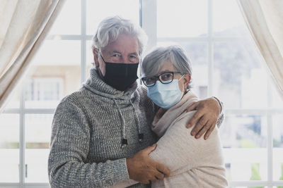 Portrait of senior couple wearing mask embracing while standing by window at home