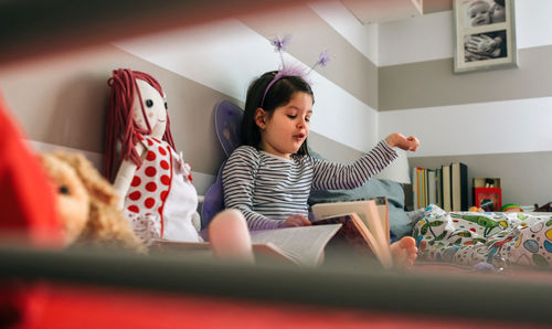 Girl reading book while sitting on bed at home