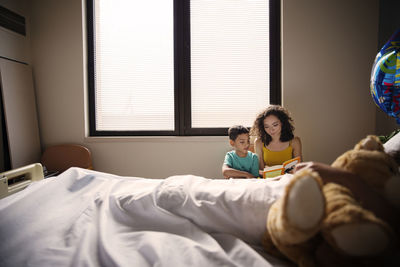 Mother and son reading book while sitting by patient in hospital ward