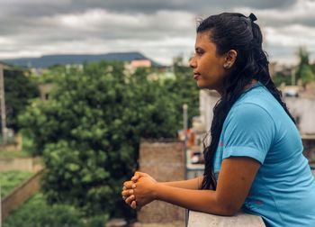 Side view of young woman looking away against trees and sky