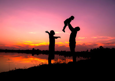 Silhouette man playing with daughter while standing by lake against sky during sunset