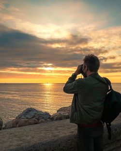 Rear view of man photographing sea against sky during sunset