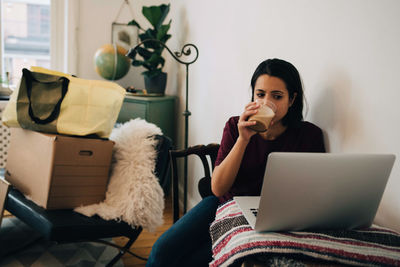 Woman drinking tea while using laptop by wall during relocation of house