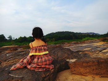 Rear view of girl sitting on rock against sky