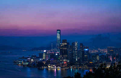 Illuminated buildings by river against sky at sunset