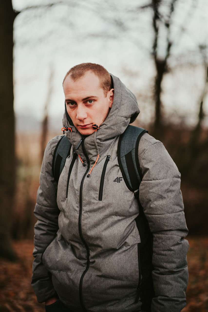 one person, looking at camera, focus on foreground, clothing, portrait, waist up, real people, lifestyles, standing, winter, young adult, leisure activity, warm clothing, day, front view, tree, jacket, nature, outdoors, scarf, hood - clothing