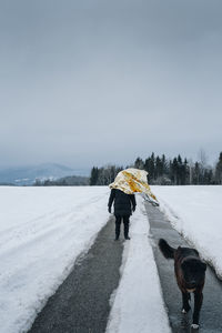 Dog walking on road by man with foil amidst snowy landscape against sky