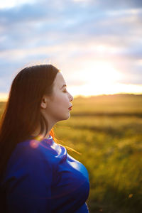 Side view of young woman looking away while standing on field against sky during sunset