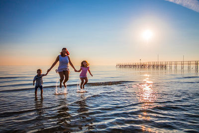 Mother holding children hands while jumping on shore at beach against sky during sunset