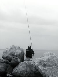 Rear view of man sitting on rock by sea against sky