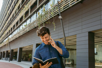 Smiling young businessman looking at diary while talking on smart phone outside office building