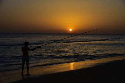 Silhouette man fishing on beach during sunset