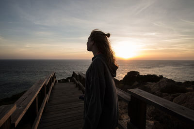 Side view of woman standing on pier at beach against sky during sunset