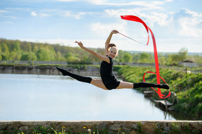 Side view of girl with ribbon jumping over retaining wall against lake