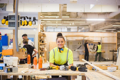 Portrait of smiling young female trainee leaning on workbench with power tools at illuminated workshop