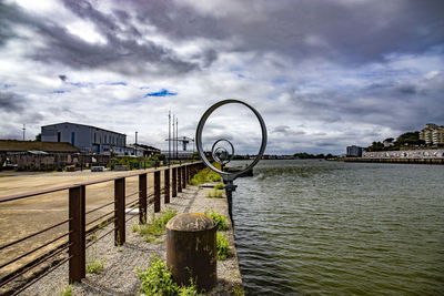 Metal railing by river in city against sky