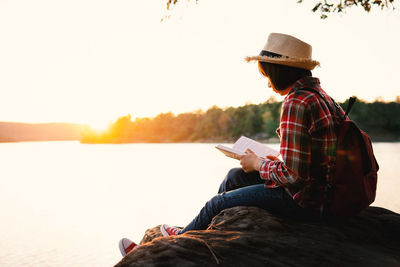 Man sitting on book by lake against sky during sunset