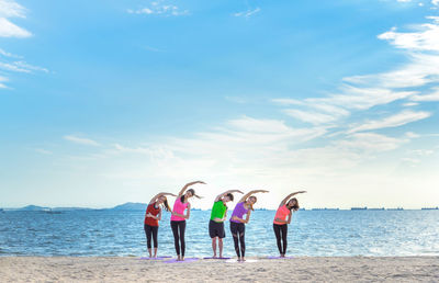 Young friends stretching arms while standing on shore at beach during sunny day