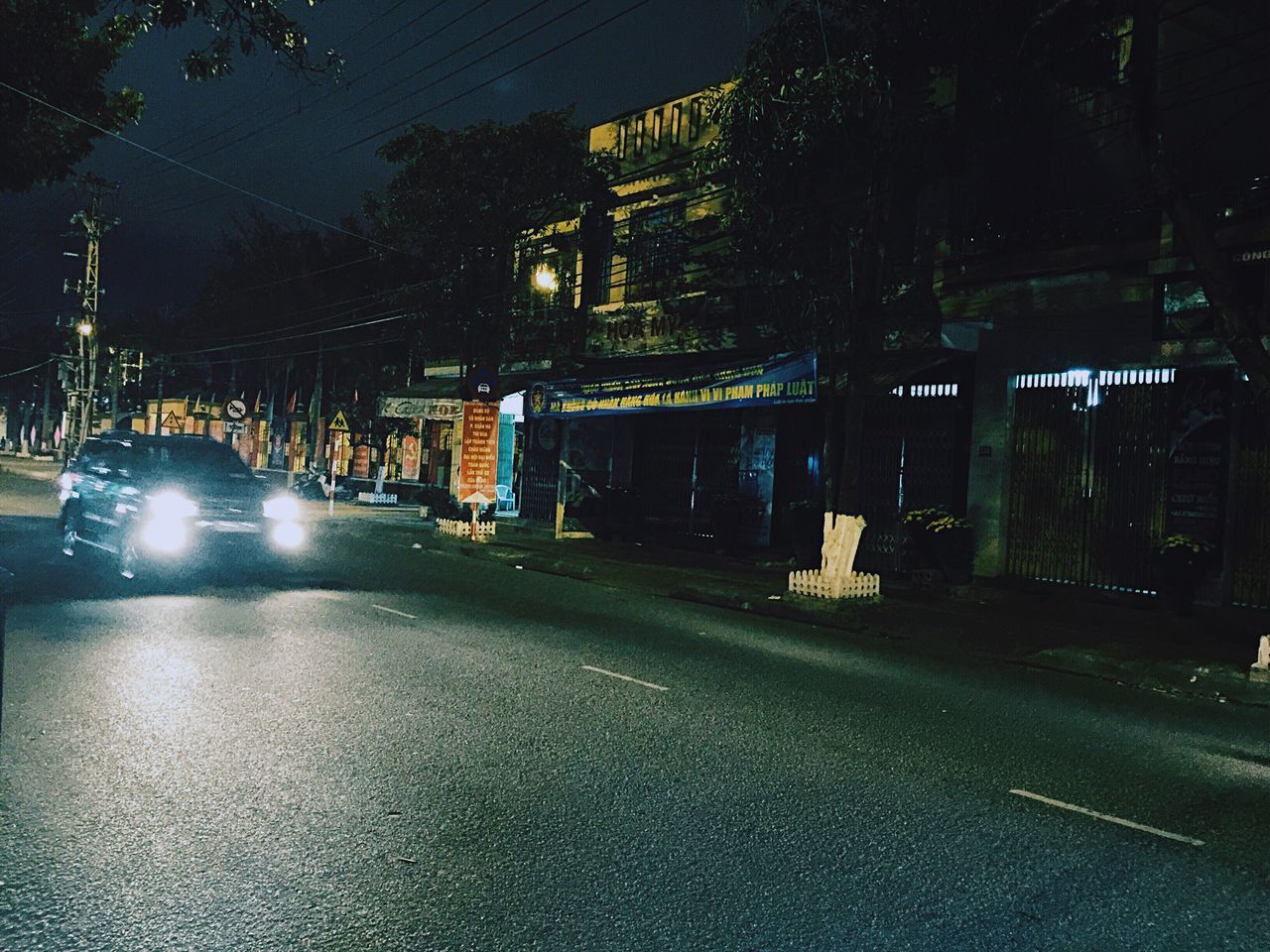 tree, the way forward, street, building exterior, night, road, illuminated, built structure, architecture, transportation, city, street light, empty, sidewalk, diminishing perspective, footpath, outdoors, road marking, vanishing point, empty road